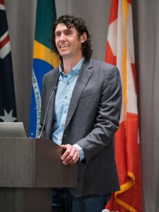 Ben Goldfarb standing at a podium in a gray suit coat and blue button down shirt.