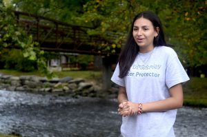 Jasmine Smith, a young woman with long dark hair, pictured with hands clasped standing next to the Oconaluftee River wearing a shirt that reads Water Protector.