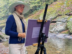 A woman in a hat stands before an art easel next to a river.