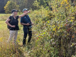 A woman wearing a backpack points out something in a fieldd to a man wearing a baseball cap.
