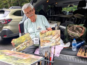 A man sitting in the back of his vehicle holding two canvas artworks.