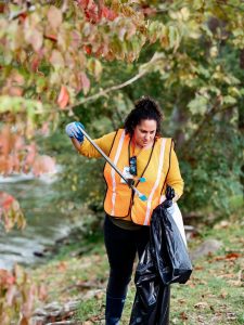 A woman wearing an orange safety vest uses grabbers to pick up trash along a river.