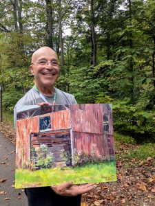 A smiling man in glasses holds an oil painting of a barn.