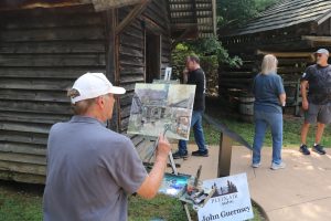 A man in a white ball cap paints on an easel next to a cabin as people walk by