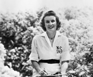Black and white photo of a woman in a white colored dress with the sleeves rolled up to her elbows and a black belt, sitting in front of some bushes.