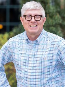 Smiling man with black rimmed glasses wearing a blu and white checkered button down shirt