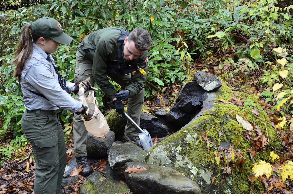 A male and female forest ranger picking up trash and shoveling a fire ring.