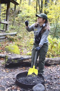 A female forest ranger wearing a baseball cap uses a yellow shovel to clean out a fire ring.