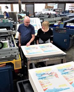 A man and woman stand next to a printing press inspecting poster reporductions.