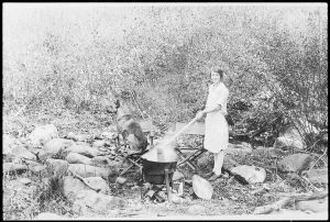 Black and white photo circa 1935 of a woman stirring a pot over an outdoor fire with a german sheppard nearby