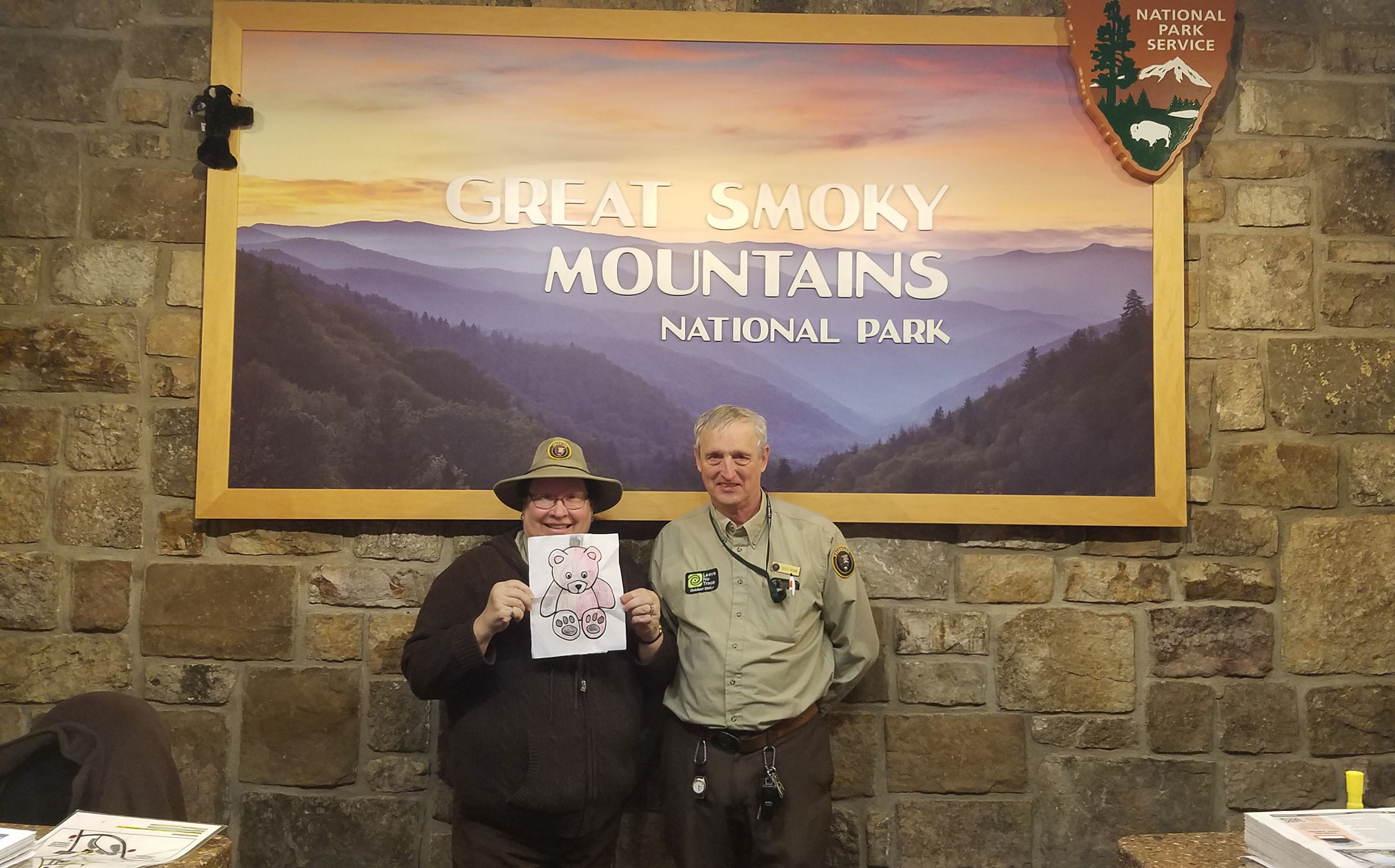 A woman, holding a coloring page, and a man in a volunteer park uniform stand beneath a GSMNP sign.