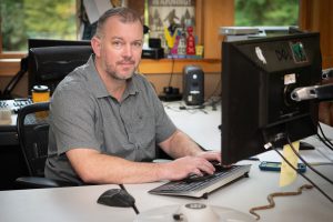 A man in a gray polo shirt sits at a computer