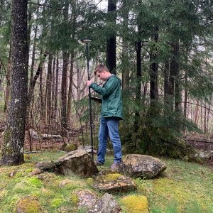 A man in a green rain jacket holding a GPS unit standing in the woods.