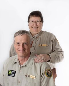 An older couple in volunteer uniforms pose in front of a white backdrop