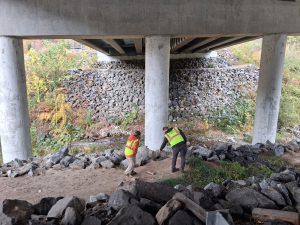 Two people in hardhats and yellow safety vests walk along a culver running below I-40