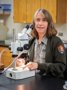 A smiling woman in an NPS ranger jacket stands in front of a microscope