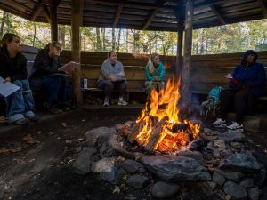 Three women sit around a bonfire inside a wooden structure
