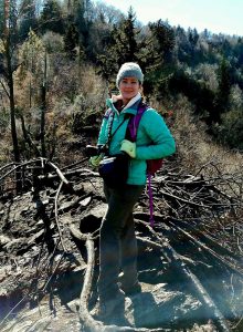 A woman in a green jacket, hat and gloves poses in the woods with a camera around her neck