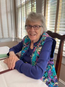 An older woman in glasses and a purple longsleeve top sits smiling at a dining room table.