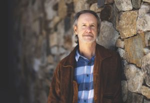 A man wearing a brown jacket over a blue and white checkered button down shirt stands against a stone wall.