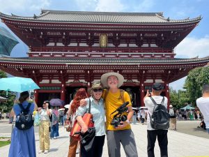 A man and woman in hats and sunglasses stand smiling on front of a Japanese building.