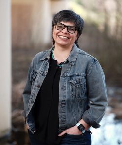 A woman with short dark hair and glasses wearing a black shirt and jean jacket smiles at the camera.
