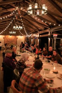 People sit at a long banquet table covered in a while table cloth with string lights hanging overhead.