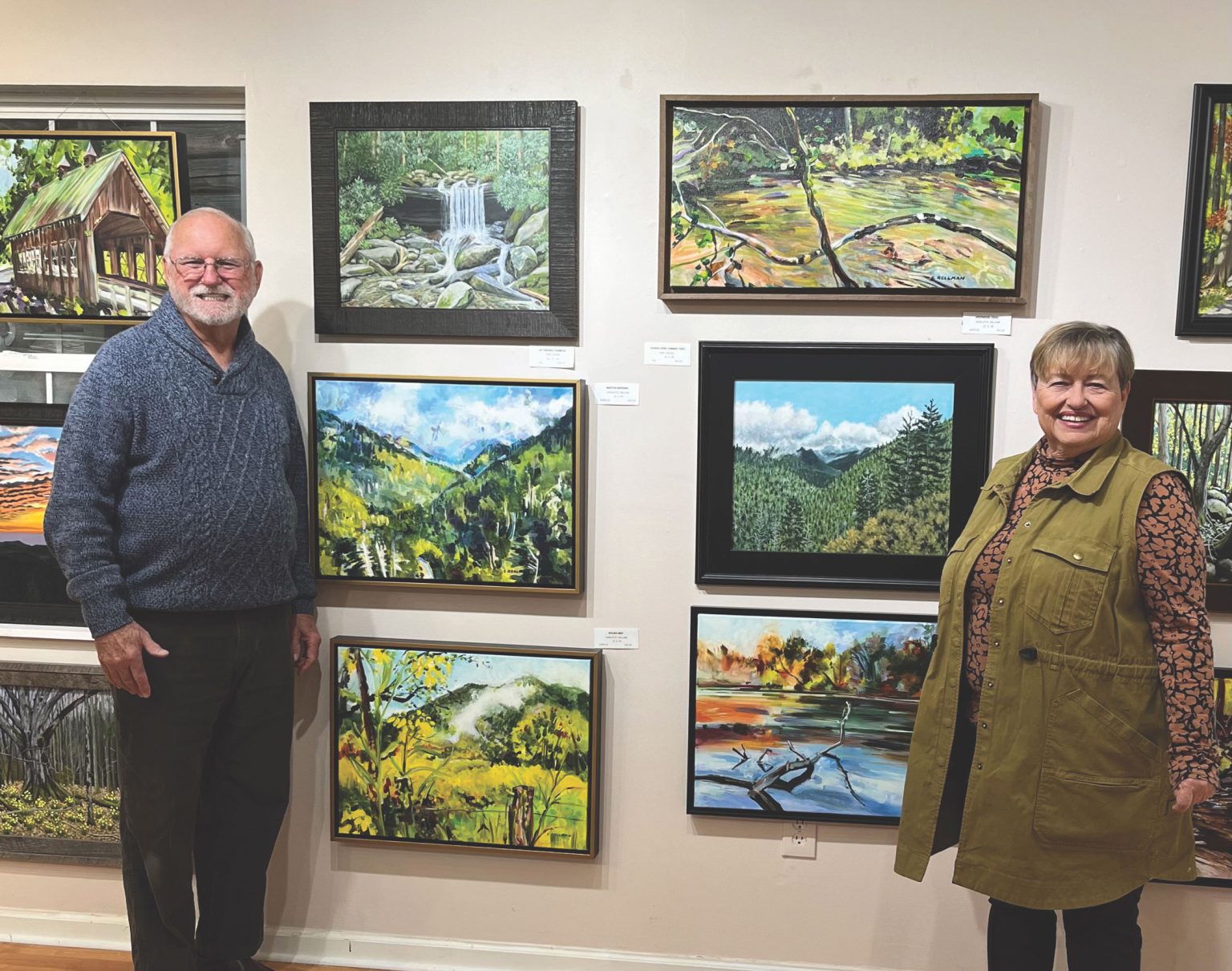 A man and woman stand next to an art gallery wall with paintings between them.