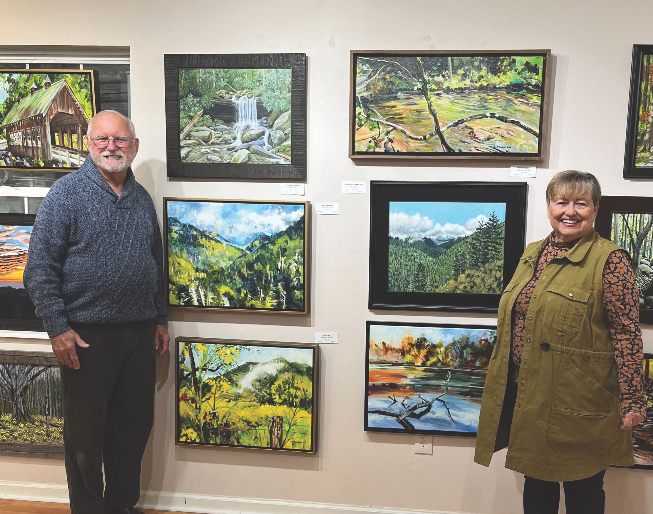 A man and woman stand next to an art gallery wall with paintings between them.