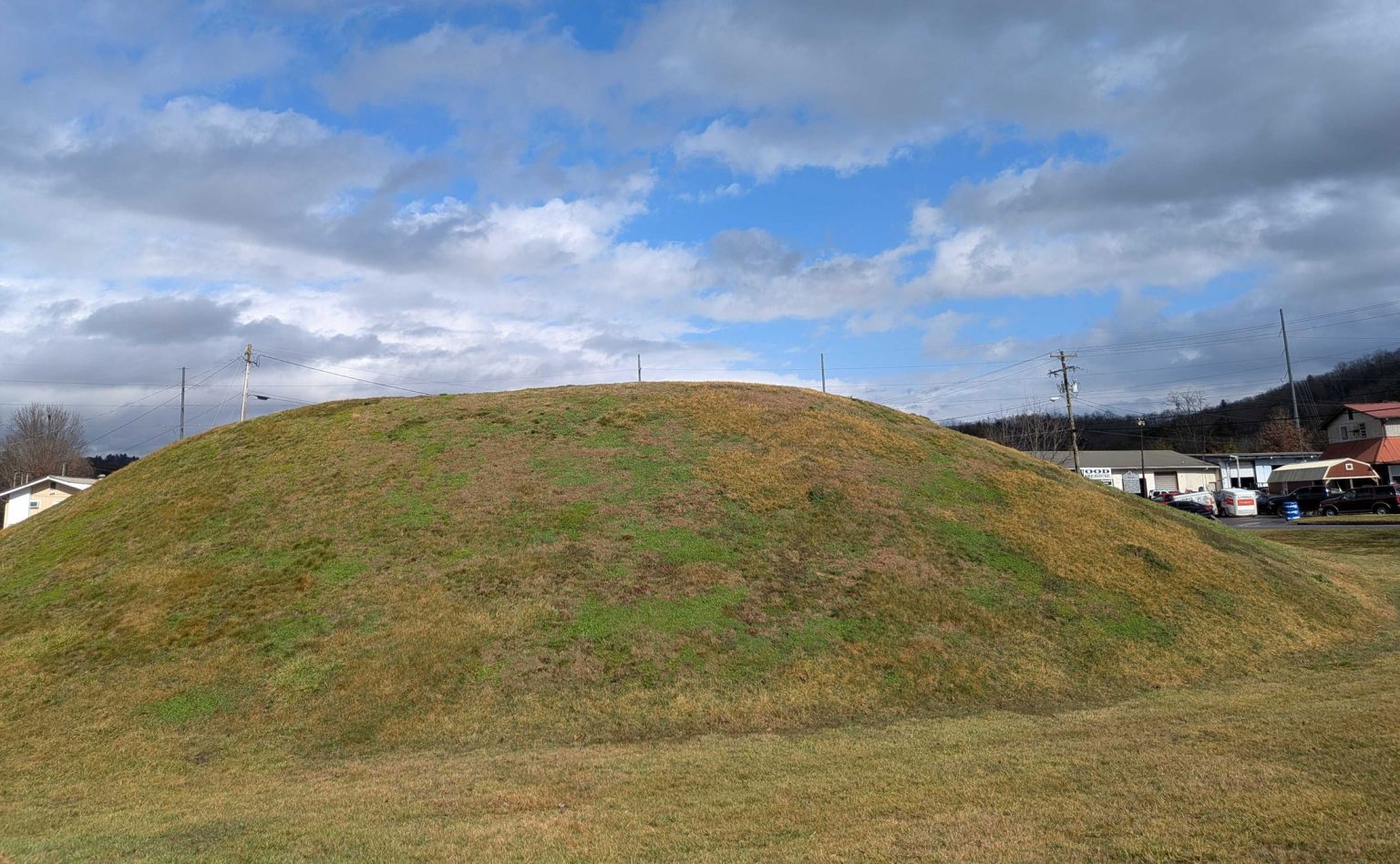A mound covered in grass beneath a cloud filed blue sky.