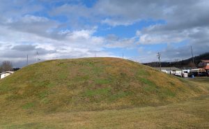 A mound covered in grass beneath a cloud filed blue sky.