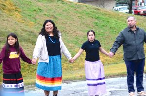 Two girls in skirts hold hands with a woman in a skirt and a man in a gray coat while performing a dance.