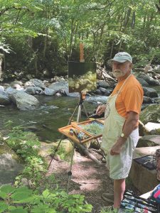A man in a baseball cap, orange shirt, khaki shorts and an apron stands next to an easel set up beside a river.