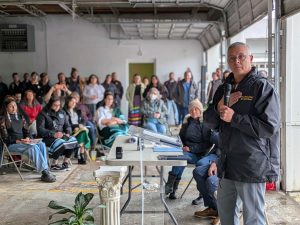 A man holds a microphone in one hand and his other hand over his heart as he speaks to a group of people inside a garage.