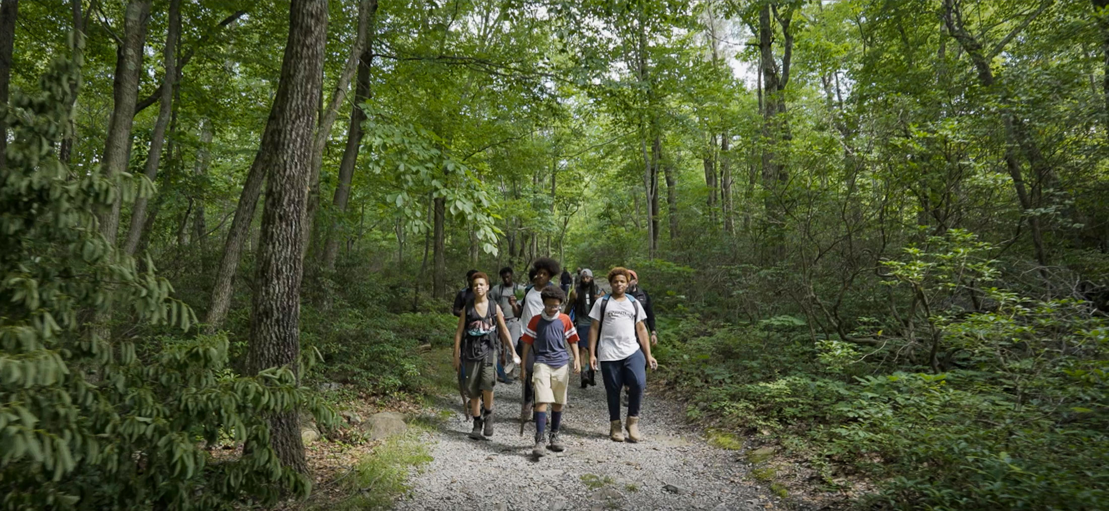 A group of black teens wearing backpacks walking towards the camera along a trail in the woods.