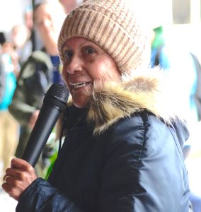 A woman in a beige winter cap and fur lined hooded jacket holding a microphone.