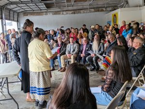 People in folding chairs watch while three standing women lead singing.