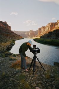 A man looks into a video camera set up alongside a river running through a canyon. 
