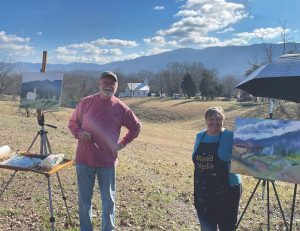 A man and woman stand next to art easels outside beneath blue sky with clouds