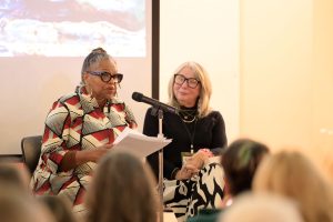 Two women sit side by side in front of an audience while one reads from a stack of papers into a microphone