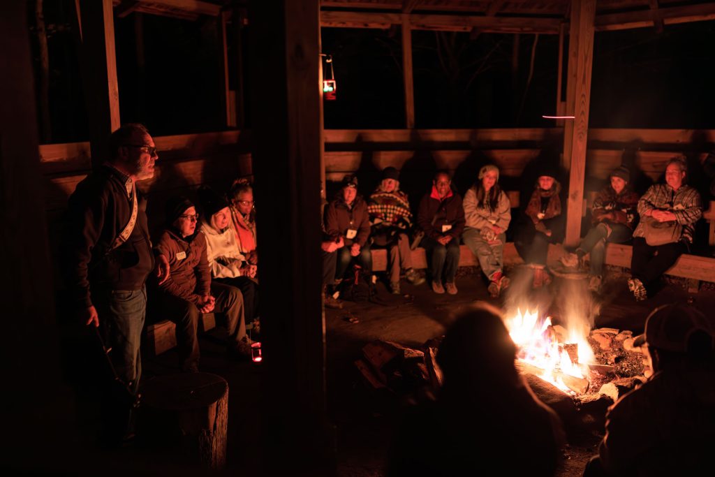 A group of people gather around a bonfire in the dark. They are front lit from the fire.
