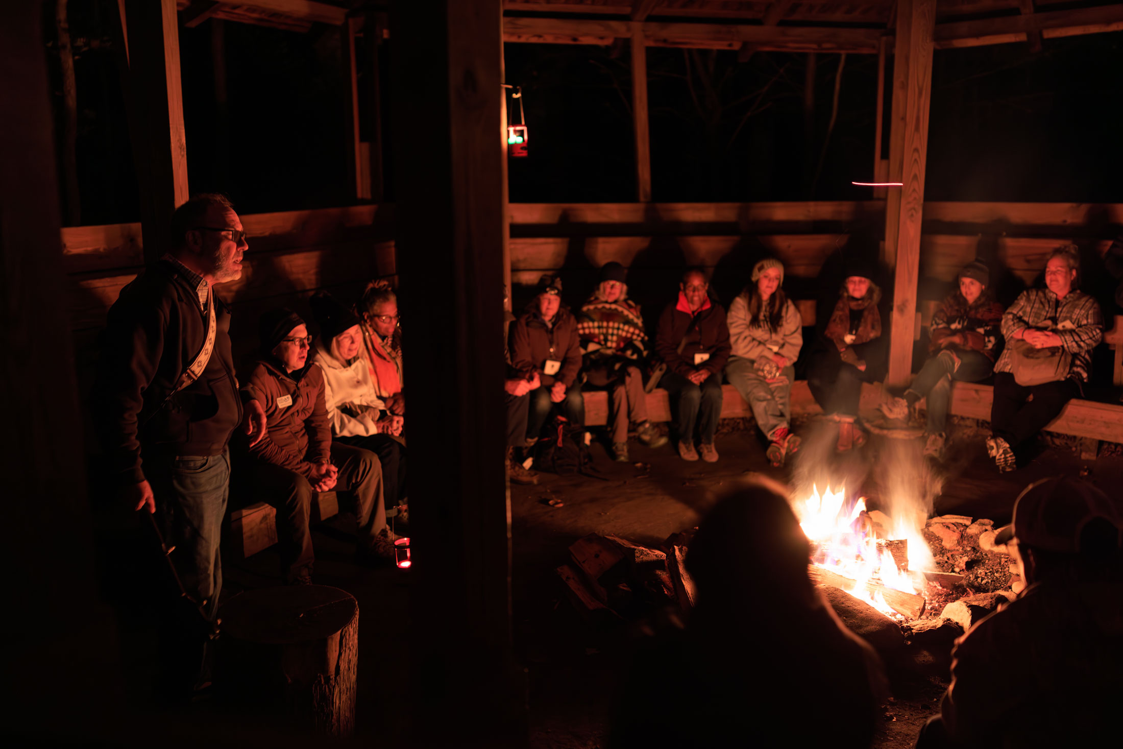 A group of people gather around a bonfire in the dark. They are front lit from the fire.