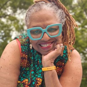 A smiling woman wearing large framed glasses holds her chin in her hand.