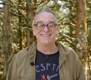 A headshot of a man with gray hair and glasses in a tan button down shirt open over a tshirt.