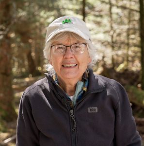 A headshot of a woman with gray hear wearing a baseball cap and glasses