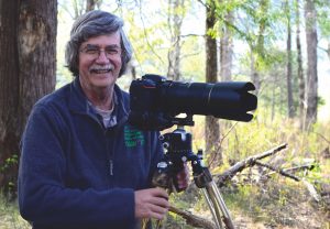 A smiling man in glasses poses next to a large lens camera.