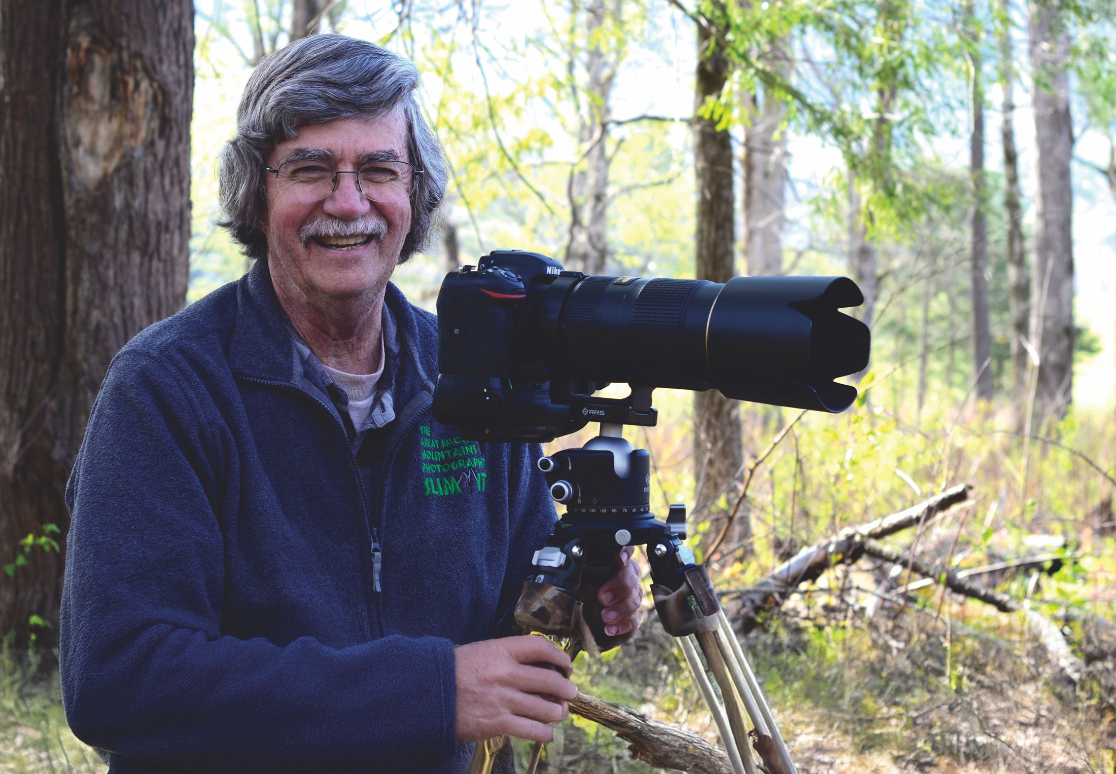 A smiling man in glasses poses next to a large lens camera.