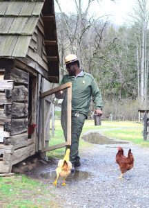 A man in a ranger uniform stands next to the open door of a chicken coop with two chickens at his feet.