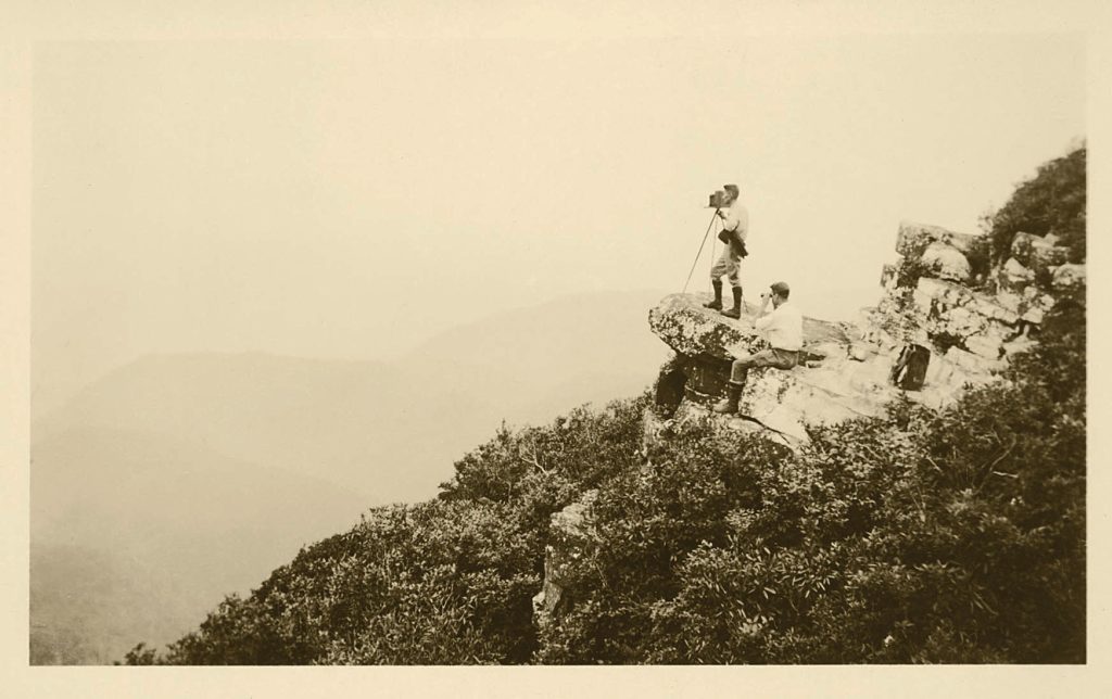 Black and white photo of two men in the distance standing on a rock with a camera