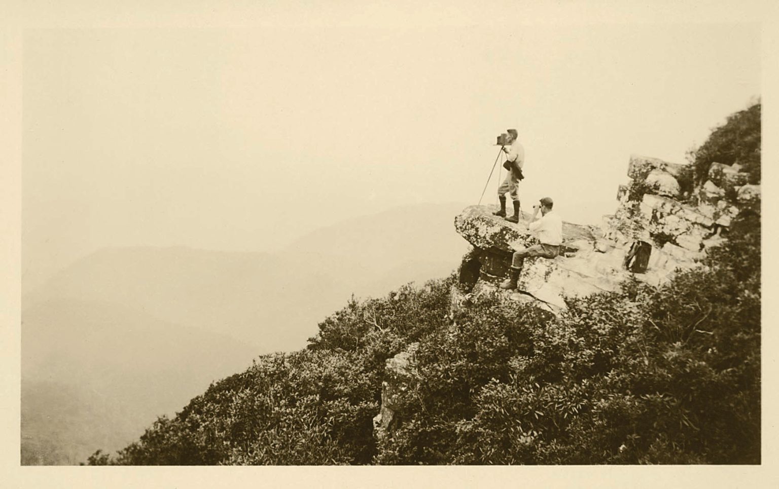 Black and white photo of two men in the distance standing on a rock with a camera